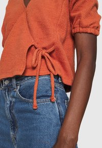 Rostfärgad kortärmad topp med texturerad yta och knytning vid midjan, tillsammans med blå högmidjade jeans.