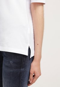 Bras droit et main d'une personne portant une chemise blanche à manches courtes et un jean bleu foncé avec une légère usure au niveau de la zone de la poche.