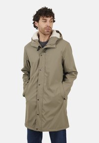 Manteau vert olive résistant à l'eau avec une capuche doublée en fausse fourrure, des boutons pression et des poches latérales, offrant une coupe décontractée et une longueur mi-cuisse.