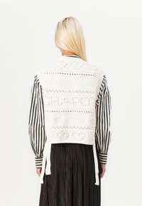 White knitted vest featuring a text pattern "CHAPOT," decorative hearts, and openwork details. Ties at the waist, worn over a striped blouse.