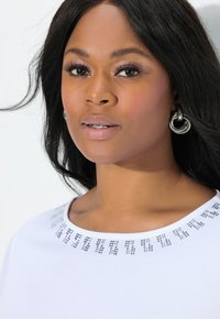 Top blanco con escote redondo que presenta tachuelas decorativas plateadas alrededor del cuello. El modelo tiene el pelo negro peinado de forma suelta y lleva pendientes de aro plateados.