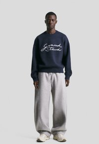 Mlad moški, oblečen v morsko modro pulover s tekstom "Grand Staud", svetlo sivo široke trenirke in bele superge, stoji pred enobarvno ozadje.