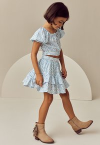 Világoskék virágmintás rövid felső puffos ujjakkal, hozzáillő, bővülő szoknyával párosítva. Bézs bokacsizmával viselve, amelyen bojtos díszítések találhatók.