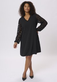 Abito nero con scollatura a V, maniche lunghe in pizzo e una silhouette fluente, abbinato a décolleté nere a punta.