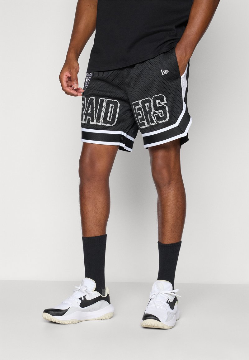 Person trägt schwarz-weiße Raiders Basketball-Shorts, schwarze Socken und weiße und schwarze Sportschuhe und steht mit einer Hand in der Tasche.