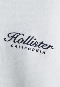 Λευκό ύφασμα με κεντημένο το κείμενο "Hollister California" σε σκούρο μπλε καλλιγραφία και με έντονα πεζά γράμματα. Μαλακή υφή, χαλαρός σχεδιασμός.