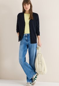 Femme portant un cardigan noir boutonné, un haut jaune, un jean bleu et des baskets blanches, tenant un sac beige souple, debout devant un mur clair.