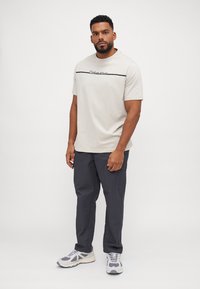 Beež crew neck t-särk musta trükiga, paarituna tumedate hallide cargo-pükstega ja hallide spordijalatsitega, millel on valged detailid.