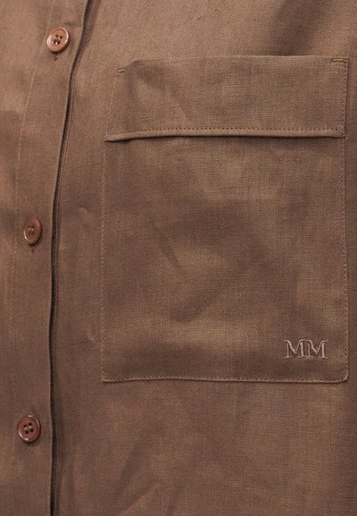 Λεπτομέρεια καφέ υφασμάτινου πουκαμίσου που δείχνει μια τσέπη με καπάκι με κέντημα "MM" και τέσσερα καφέ κουμπιά κατά μήκος της μπροστινής πατιλέτας.