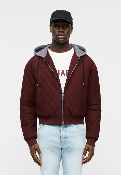 Homme portant une casquette noire, une veste matelassée bordeaux zippée avec une capuche grise, un T-shirt blanc imprimé et un jean bleu clair, se tenant devant un fond blanc.