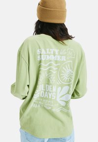 Zielona koszula z długim rękawem, wykonana z miękkiego materiału, z białym graficznym napisem i tropikalnymi wzorami, w tym falami i liśćmi na plecach.