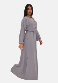 Robe longue grise en tissu léger. Présente un décolleté en V profond, des manches longues et des boutons sur le devant. Texture lisse avec un design fluide.