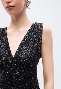 Μαύρο φόρεμα με παγιέτες, με βαθιά V λαιμόκοψη και φαρδιές τιράντες, που αναδεικνύει μια υφασμάτινη, λαμπερή επιφάνεια με εφαρμοστή σιλουέτα.