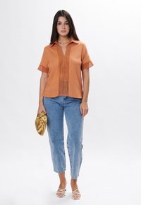 Rustrød skjorte med krave, halværmer og lodrette udkæringer. Sammen med lyseblå jeans og en gylden clutch. Hvide strop sandaler.