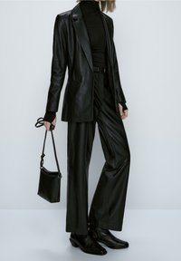 Zwarte leren blazer over een zwarte coltrui, gecombineerd met wijdvallende leren broeken en enkellaarsjes. Geaccentueerd met een kleine zwarte handtas.