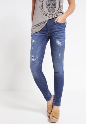 Femme portant un jean skinny bleu usé associé à des sandales compensées beiges à bout ouvert et un haut gris sans manches avec un motif de crâne.