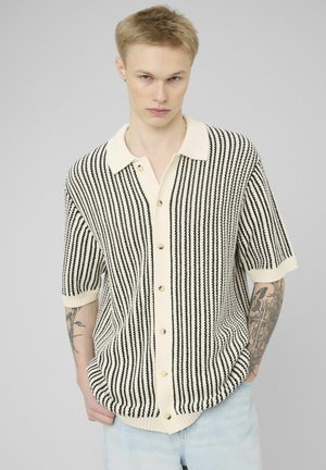 Jeune personne aux cheveux blonds courts portant une chemise à manches courtes à rayures verticales noires et blanches, un jean clair et des tatouages visibles sur les bras.