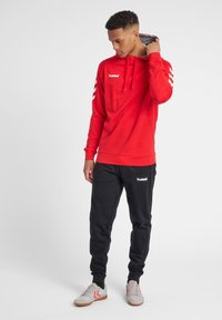 Felpa rossa con cappuccio regolabile, logo bianco e dettagli a righe sulle maniche, abbinata a pantaloni jogger neri con logo bianco.