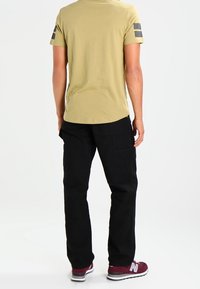 Olijfgroene t-shirt met korte mouwen en zwarte strepen op de mouwen, gecombineerd met zwarte cargo pants en kastanjebruine sport schoenen.
