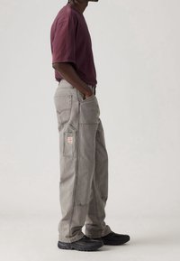 Pantalones cargo grises con múltiples bolsillos, detalles cosidos y un parche de marca. Combinados con una camiseta ajustada color burdeos y zapatillas negras.