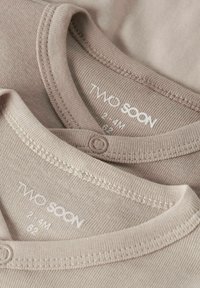 Deux bodysuits en coton beige avec une texture côtelée, présentant des fermetures à pression, des encolures arrondies et des étiquettes de marque affichant "TWO SOON."