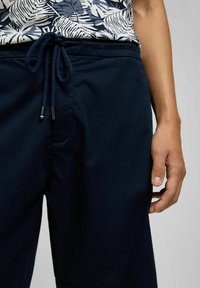 Marineblaue Shorts mit elastischem Bund und Kordelzug, ausgestattet mit einer Vorderasche und weichem Stoff, kombiniert mit einem weißen, floral gemusterten Hemd.