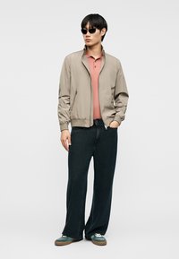 Άνδρας που φοράει μπεζ μπουφάν bomber πάνω από ροζ πόλο πουκάμισο, μαύρο τζιν με φαρδιά πόδια, πράσινα αθλητικά παπούτσια και μαύρα γυαλιά ηλίου, στέκεται με το ένα χέρι στην τσέπη.