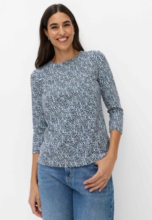 Donna sorridente che indossa una camicia a maniche lunghe blu e bianca con fantasia e jeans blu, in piedi con una mano in tasca su sfondo bianco.