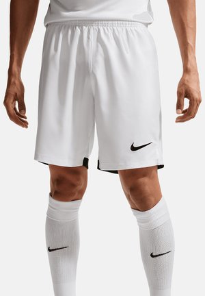 Mann trägt weiße Nike Fußballshorts und weiße kniehohe Socken mit schwarzen Nike Swoosh-Logos auf einfachem Hintergrund.