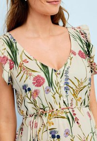 Bloemjurk met een v-hals, korte gerimpelde mouwen en een aansluitende tailleband. Heeft kleurrijke bloemenpatronen op een crème achtergrond.
