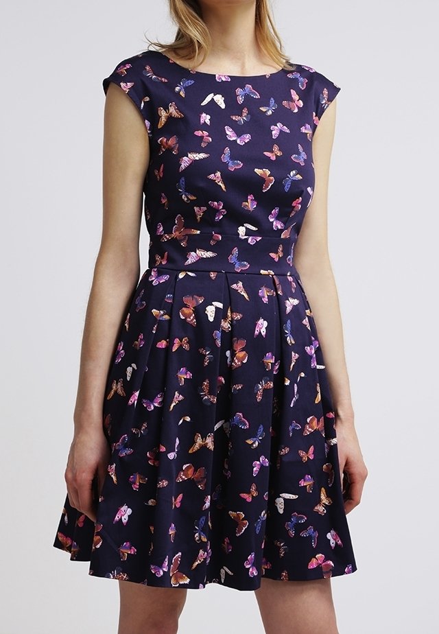 Kvinna iklädd en ärmlös marinblå klänning med utsvängd kjol, dekorerad med flerfärgade fjärilsmotiv.