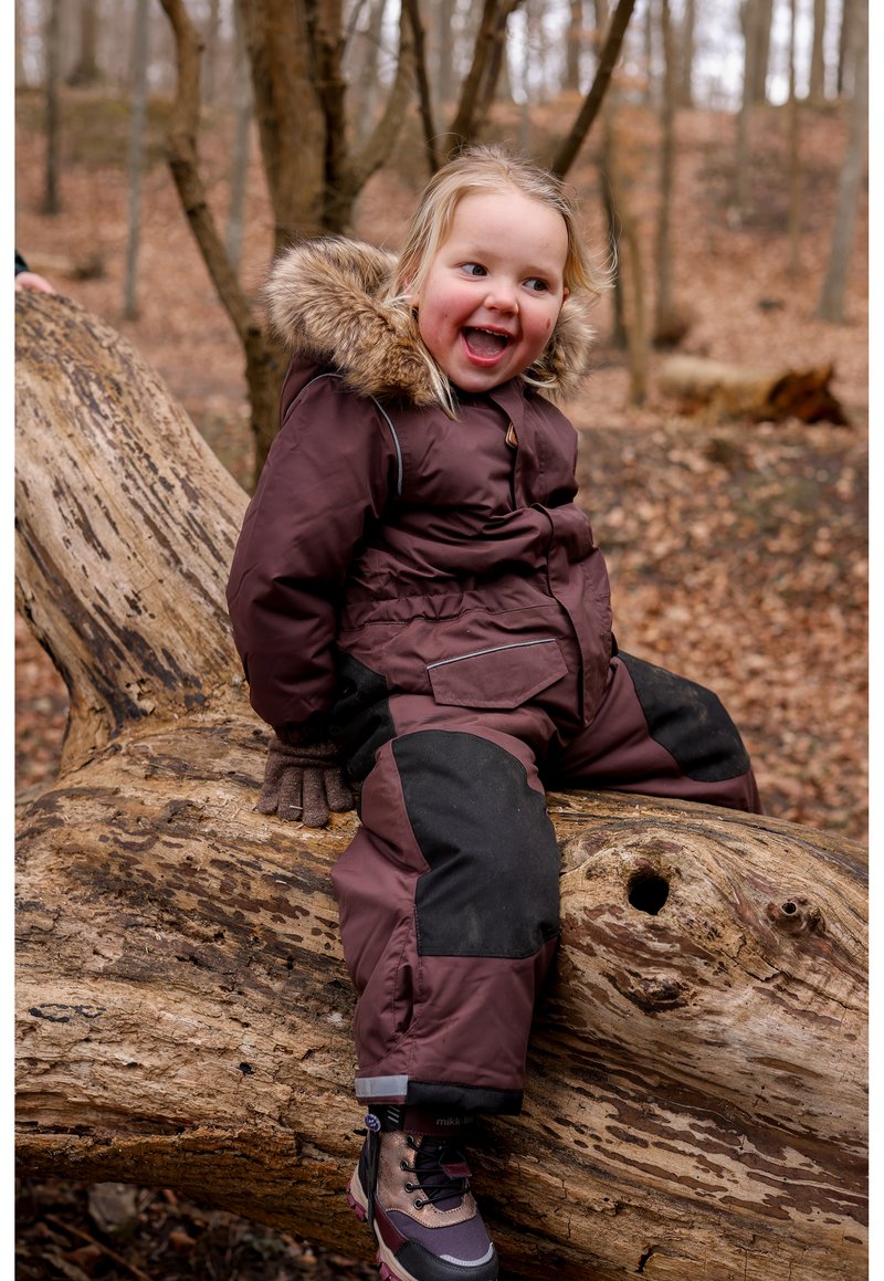 Zateplená bordó kombinéza na sníh s kapucí lemovanou kožešinou, černými doplňky, pevnou konstrukcí a rukavicemi, kterou nosí dítě sedící na pařezu.
