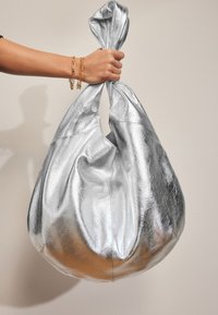 Ezüst fémes táska, texturált anyagból, laza formával és csomózott fogantyúval, egy kézben, amelyen arany lánc karkötők vannak.