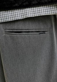 Poche arrière d'un pantalon gris avec une chemise à carreaux noir et blanc rentrée, montrant une petite étiquette Jack & Jones sur la poche.