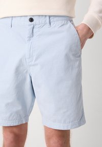 Calções de algodão azul claro com um corte descontraído, apresentando um fecho frontal com botão, bolsos laterais e detalhes de bainha cosida.