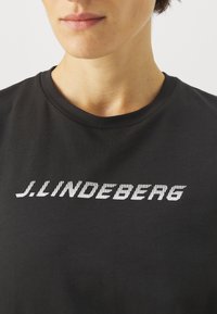 Černé bavlněné tričko s bílým pruhovaným logem "J.LINDEBERG" na přední straně. Design s kulatým výstřihem, hladká textura.