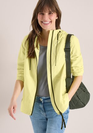Femme souriante portant une veste à capuche jaune clair, un t-shirt gris, un jean bleu, et un sac bandoulière matelassé vert foncé sur fond clair.