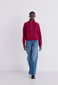 Pull en polaire rouge avec un zip jusqu'à mi-hauteur, poche avant, et manches longues, associé à un jean large bleu clair et des chaussures noires.