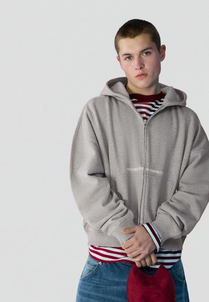 Junger Mann trägt eine graue Kapuzenjacke mit Reißverschluss über einem rot-weiß gestreiften Hemd, hält eine rote Mütze und steht vor einem schlichten hellen Hintergrund.