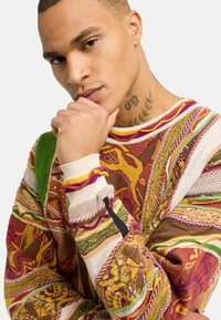 Pull multicolore présentant des motifs complexes, texture en tricot et poignets côtelés. Inclut des accents verts et jaunes avec un design unique.