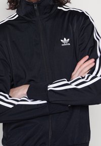 Czarna kurtka sportowa na suwak z białymi trzema paskami na rękawach, z wyhaftowanym logo Adidas na lewej piersi.