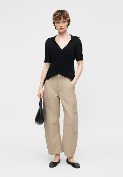 Νεαρό άτομο που φορά μαύρο μπλουζάκι με κοντά μανίκια, μπεζ παντελόνι cargo φαρδύ, μαύρα φλατ παπούτσια, κρατώντας μικρή μαύρη τσάντα, στέκεται μπροστά σε μονόχρωμο φόντο.