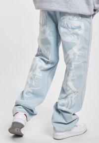 Vaqueros de mezclilla azul claro con gráficos de dragones blancos, ajuste relajado y dobladillos remangados. Incluyen bolsillos tradicionales y zapatillas blancas.