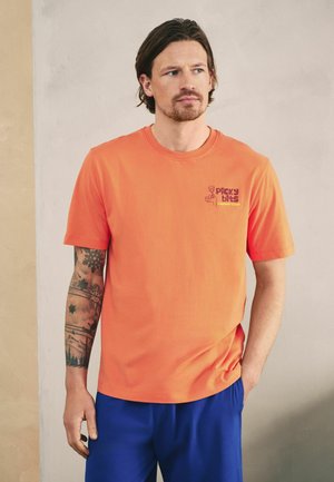 Bărbat cu tatuaje, purtând un tricou portocaliu cu inscripția „Picky Bits Supper Club” și pantaloni albaștri, stând cu o mână în buzunar, sprijinit de un perete simplu.