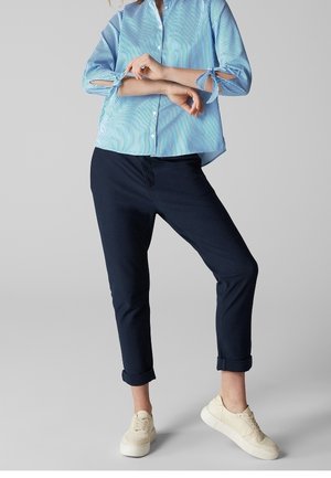 Persona de pie con los brazos cruzados, vistiendo una blusa azul a rayas, pantalones oscuros remangados y zapatillas blancas, frente a un fondo liso.