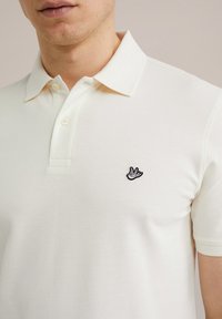 Krémová polo košile z texturované látky, s černě vyšitým logem ptáka na levém prsu a klasickým límečkem se třemi knoflíky.