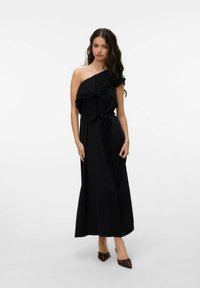 Kobieta w czarnej, jedno-szerokiej sukience maxi z falbanką i przewiązanym w pasie paskiem, nosząca brązowe czółenka z oczkowymi kokardkami.