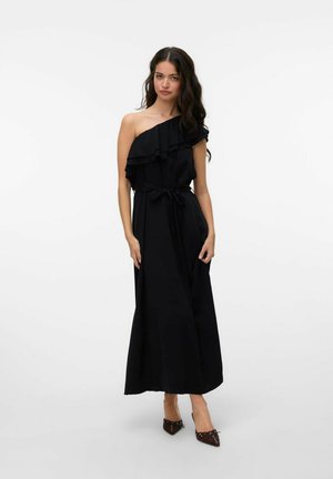 Vestido negro de un solo hombro con detalles de volantes, cintura ceñida, falda fluida y tejido suave. Usado con tacones marrones de punta.