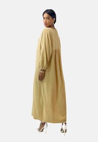 Robe longue et fluide en tissu beige doux avec une surface texturée, présentant un col haut et une coupe ample, agrémentée de manches longues.