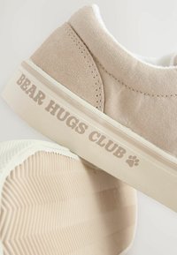 Beige Leinenschuhe mit einem flauschigen Innenfutter, ausgestattet mit einer Gummisohle und dem Schriftzug "BEAR HUGS CLUB" an der Seite. Dezente Textur, niedrig geschnittenes Design.